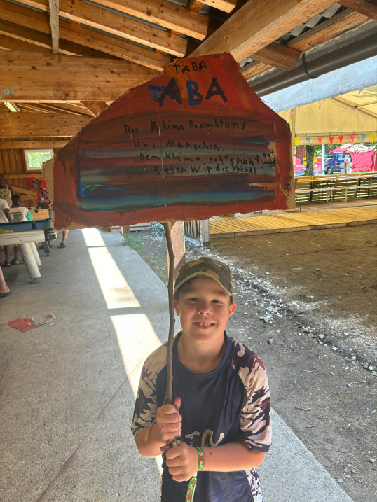 Ein Kind steht in einer Holzhalle und hält ein selbst bemaltes Pappschild an einem Stock. Das Schild hat bunte Farben und einen Text zum Thema Klima.