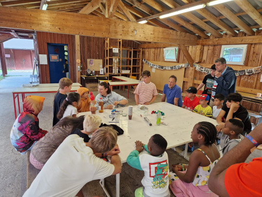Eine große Gruppe von Kindern und Erwachsenen sitzt an einem langen Tisch in einer Halle. Auf dem Tisch liegen Bastel- und Experimentmaterialien.