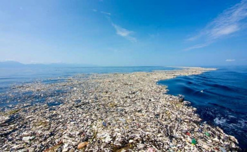 Das Foto zeigt die Wasseroberfläche im Pazifik, die großflächig mit schwimmendem Müll bedeckt ist. Plastikflaschen, Tüten und andere Abfälle treiben dicht an dicht und bilden einen grauen Teppich, der bis zum Horizont reicht. Rechts im Bild ist ein schmaler Streifen klaren, blauen Wassers zu sehen, während sich darüber der blaue Himmel mit einigen Wolken erstreckt.