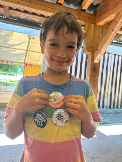 Ein Kind hat drei bunte Buttons am T-Shirt. Die Buttons sind selbst gestaltet und zeigen verschiedene Bilder und Schriftzüge: Die Schildkröte Taba, den Schriftzug "Retten wir die Welt" und die Beschriftung "Klimafüxe" auf buntem Hintergrund.
