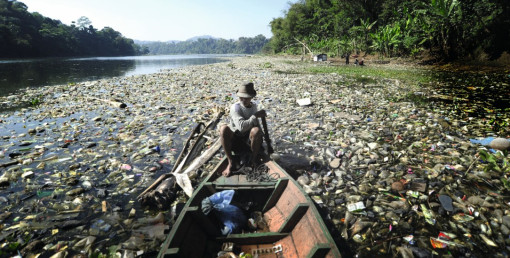 Ein Mann sitzt in einem kleinen Holzboot auf einem Fluss, der stark mit Plastikmüll, Flaschen und anderem Abfall verschmutzt ist. Das Wasser ist kaum sichtbar, weil es von Abfall bedeckt ist. Am Ufer wachsen Bäume und Pflanzen, im Hintergrund sind einige Menschen und eine kleine Hütte zu sehen.