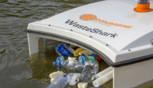 Ein kleines, weißes, schwimmendes Gerät mit der Aufschrift „WasteShark“ sammelt Plastikmüll und Flaschen von der Wasseroberfläche ein. Es schwimmt auf einem Gewässer, während verschiedene Plastikflaschen und Abfallstücke vorne in das Gerät hineingezogen werden.