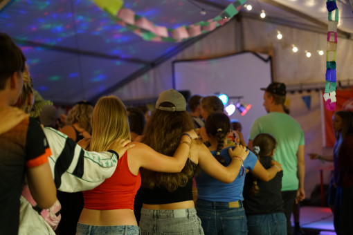 Viele Menschen stehen im Zelt und haben die Arme umeinander gelegt. Bunte Lichter und Papierketten hängen über der Gruppe.
