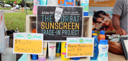 Ein Marktstand in Hawaii zeigt viele Sonnencremeflaschen verschiedener Marken und Größen. Im Zentrum hängt ein Schild mit dem Aufdruck: „The Great Sunscreen Trade-In Project – Hawaii says NO to toxic sunscreens“. Links und rechts stehen handgeschriebene Pappschilder: „$3 off Trade Ins!“ und „These products will be illegal in 2021“. Im Hintergrund ist ein Plakat mit einer Frau und einem Kind zu sehen. Der Stand informiert über das Verbot bestimmter Sonnencremes auf Hawaii.