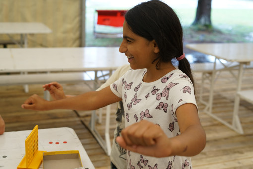 Ein Kind steht an einem Tisch und spielt ein kleines Brettspiel. Es hält die Arme ausgestreckt. Auf dem Tisch liegt ein gelbes Spiel mit roten Spielsteinen.