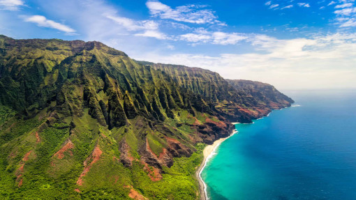 Ein Luftbild zeigt die Na-Pali-Küste auf Hawaii. Steile, grün bewachsene Felswände mit markanten Schluchten fallen direkt zum Meer hin ab. Am Fuß der Klippen liegt ein schmaler, heller Sandstrand. Davor erstreckt sich türkisfarbenes Wasser, das in der Ferne in tiefes Blau übergeht. Über der Landschaft liegt ein leicht bewölkter, blauer Himmel.
