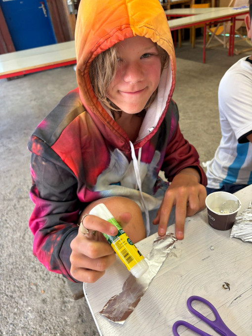 Ein Kind sitzt an einem Tisch und bastelt. Es streicht Kleber auf ein Stück Alufolie. Auf dem Tisch liegen Schere, Becher und weitere Bastelmaterialien.