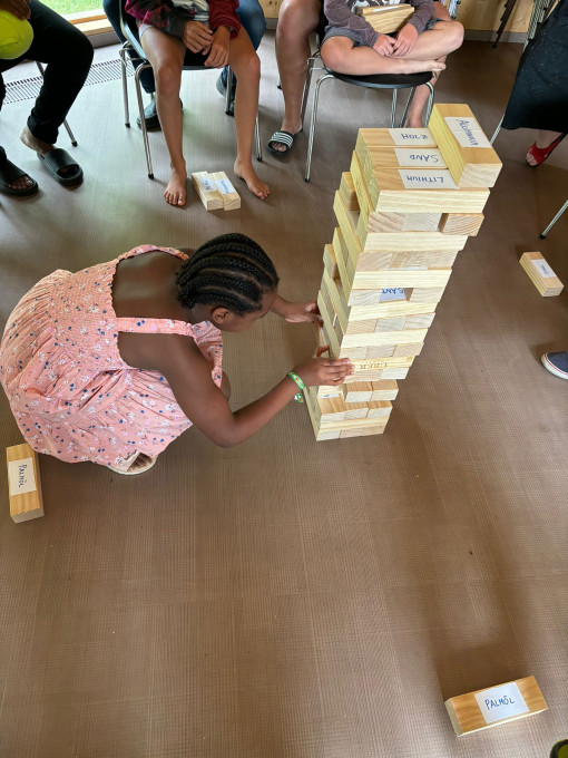 Ein Kind kniet am Boden und spielt Jenga mit großen Holzklötzen. Andere Kinder sitzen im Kreis darum. Auf den Jenga-Steinen stehen Wörter: Holz, Sand, Lithium, Aluminium
