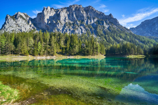 Man sieht einen klaren, grün-türkisen See in den Bergen. Das Wasser ist so durchsichtig, dass man den Boden sehen kann. Rund um den See wachsen viele grüne Nadelbäume. Dahinter ragen hohe, graue Felsen steil in den Himmel. Der Himmel ist blau mit ein paar weißen Wolken. Die Stimmung wirkt hell, freundlich und ruhig.