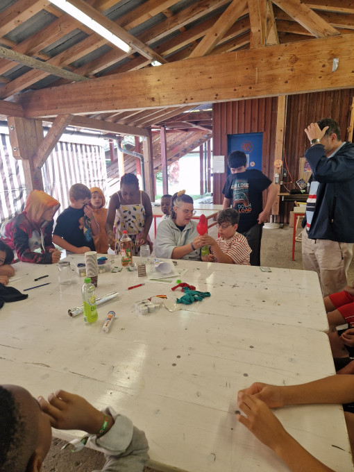 Mehrere Kinder sitzen an einem Tisch in einer Holzhalle. Auf dem Tisch stehen Materialien für ein Experiment, zum Beispiel Flaschen, Becher und Folie. Eine erwachsene Person erklärt etwas.